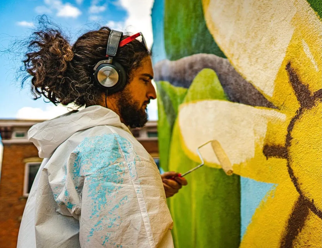 Jason Al Ghussein, wearing headphones and a painter's coverall, painting a yellow orange blossom